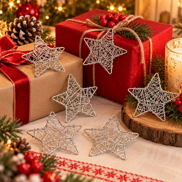 Five silver wire star-shaped ornaments arranged on a festive Christmas table with wrapped gifts, pine branches, red berries, and a lit candle in the background.