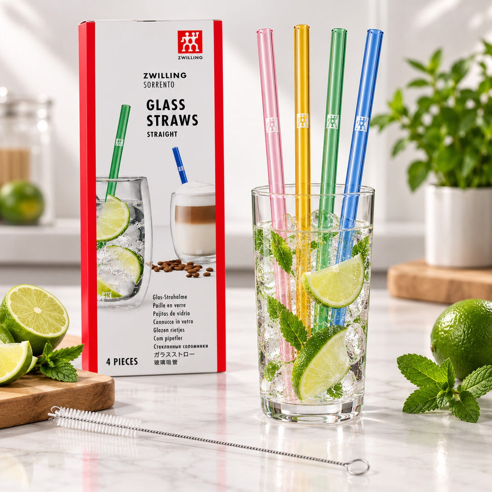 Four colored glass straws in pink, yellow, green, and blue are shown in a clear drinking glass with lime slices and mint, alongside their product packaging box and a metal straw cleaning tool on a marble surface.