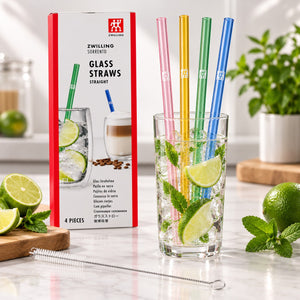 Four colored glass straws in pink, yellow, green, and blue are shown in a clear drinking glass with lime slices and mint, alongside their product packaging box and a metal straw cleaning tool on a marble surface.