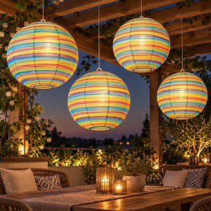 Four glowing spherical paper lanterns with horizontal rainbow stripes hang from a wooden pergola at dusk, surrounding an outdoor patio table with candles and seating.