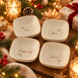 Four small square white ceramic bowls with rounded corners, decorated with gold borders and gold script words reading Joy, Cheer, Happiness, and Shine, arranged on a wooden tray with holiday lights, pine branches, ornaments, and a wrapped gift box.