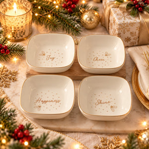 Four square white ceramic bowls with gold trim, arranged in a 2x2 grid on a wooden board. Each bowl is printed with a gold bee, small gold dots, and a holiday-themed word: 'Joy', 'Cheer', 'Happiness', and 'Shine'.