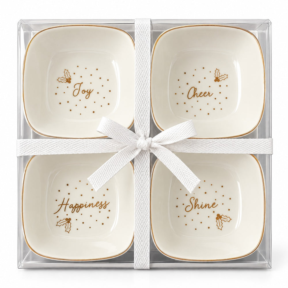 Four small square white ceramic bowls with rounded corners, decorated with gold borders and gold script words reading Joy, Cheer, Happiness, and Shine, arranged on a wooden tray with holiday lights, pine branches, ornaments, and a wrapped gift box.