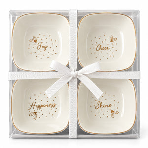 A 4-piece ceramic appetizer or dessert server set in a clear plastic gift box, tied with a white ribbon. Each square white ceramic piece has a gold rim, gold dotted border, and a gold script word (Joy, Cheer, Happiness, Shine) with a small gold leaf illustration.
