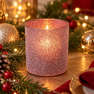 A pink glitter candle holder candle lit on a tabletop surrounded by Christmas decorations including pine branches, red berries, a pinecone, and gold ornaments.