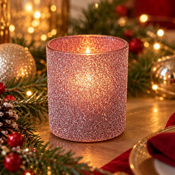 A pink glitter candle holder candle lit on a tabletop surrounded by Christmas decorations including pine branches, red berries, a pinecone, and gold ornaments.