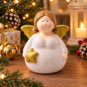 A round white angel figurine with gold glitter wings and a gold star accent, displayed on a wooden table with a warm holiday background featuring pine branches, a candle, and a gift box.