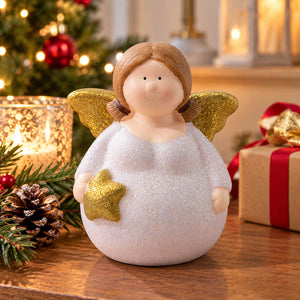 A Christmas angel figurine with a glittery white dress, brown hair, and gold wings, styled on a wooden table with a lit candle, pinecone, and wrapped gift in a festive holiday setting.