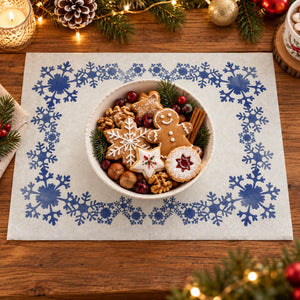 A round white placemat with a blue snowflake border pattern, styled on a wooden table with holiday decorations and a bowl of gingerbread cookies in the center.