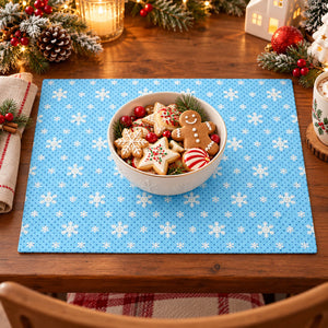 A square blue placemat with a white snowflake pattern is set on a wooden dining table, styled with a white bowl of Christmas cookies in the center and holiday decorations around the edges.