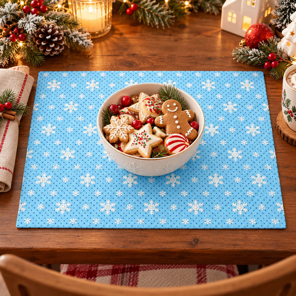 A square blue placemat with a white snowflake pattern is set on a wooden dining table, styled with a white bowl of Christmas cookies in the center and holiday decorations around the edges.