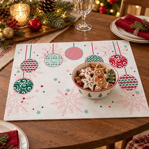 A rectangular Christmas-themed placemat on a wooden dining table, featuring a white background with red and green hanging ornaments, snowflakes, and a gingerbread man motif. A bowl of cookies sits on the placemat, with plates, candles, pinecones, and holiday decorations in the background.