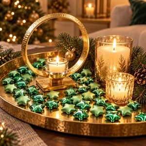 A festive holiday tabletop arrangement featuring a hammered gold tray with a large gold ring holder, lit white pillar candles in glass holders, and many shiny green star-shaped ornaments. Pine branches, a pinecone, and blurred Christmas tree lights create a warm seasonal background.