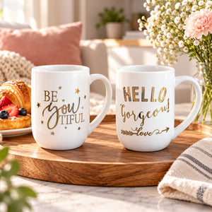 Two white ceramic mugs with gold lettering displayed on a wooden tray in a cozy kitchen setting. One mug reads 'BE YOU TIFUL' with gold stars, and the other reads 'HELLO gorgeous' with gold script and arrow details.