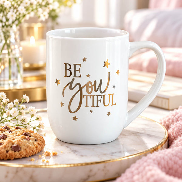 A white ceramic mug with the phrase 'BE you TIFUL' in gold script and star accents, styled on a marble tray with a cookie, flowers, and a candle in a cozy indoor setting.