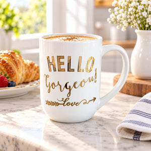 A white ceramic coffee mug with gold foil lettering that reads 'HELLO Gorgeous! love' sits on a marble countertop in a bright kitchen setting, with a croissant, flowers, and sunlight in the background.