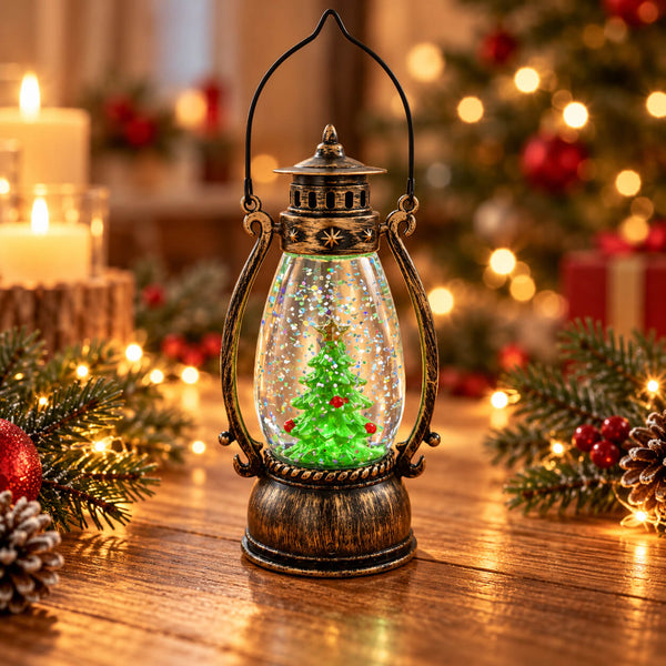A decorative lantern-shaped snow globe sits on a wooden table surrounded by Christmas greenery, pinecones, and warm string lights. The lantern has an antique bronze finish and contains a miniature glowing green Christmas tree with red ornaments inside clear snow-like spheres.