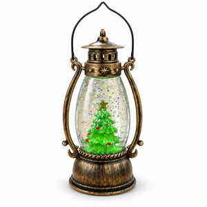 A decorative lantern-shaped holiday ornament with an antique bronze metal frame and handle, featuring a clear glass body containing a green Christmas tree with snow-like glitter inside, shown on a white background.
