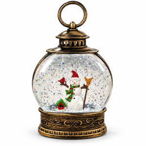 A decorative lantern-shaped snow globe with an antique bronze metal frame, featuring a miniature snowman, a small birdhouse, and glittery white internal snow inside a clear glass sphere, shown on a plain white background.