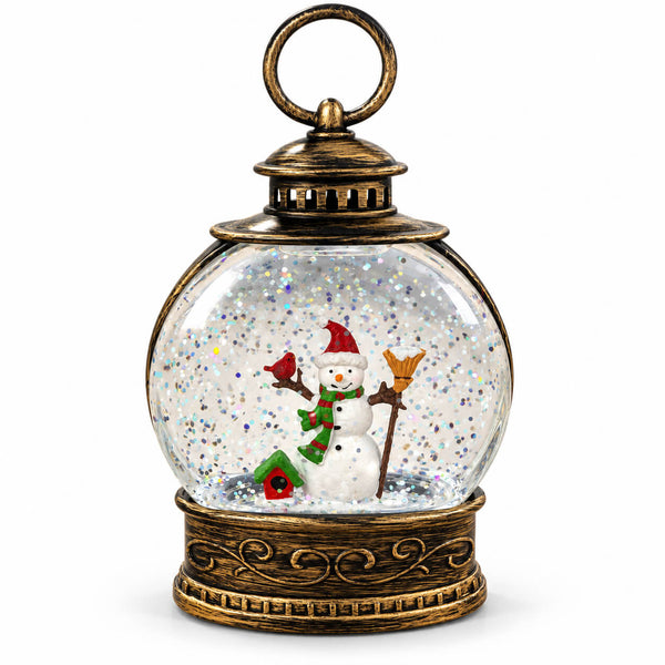 A decorative lantern-shaped snow globe with an antique bronze metal frame, featuring a miniature snowman, a small birdhouse, and glittery white internal snow inside a clear glass sphere, shown on a plain white background.