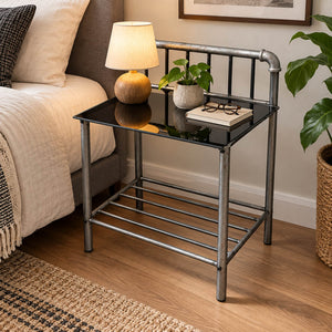 Industrial-style metal end table with a black glass top, shown beside a bed in a bedroom setting. The table has a galvanized steel frame, a lower shelf, and decorative items including a lamp, plant, and books on the tabletop.
