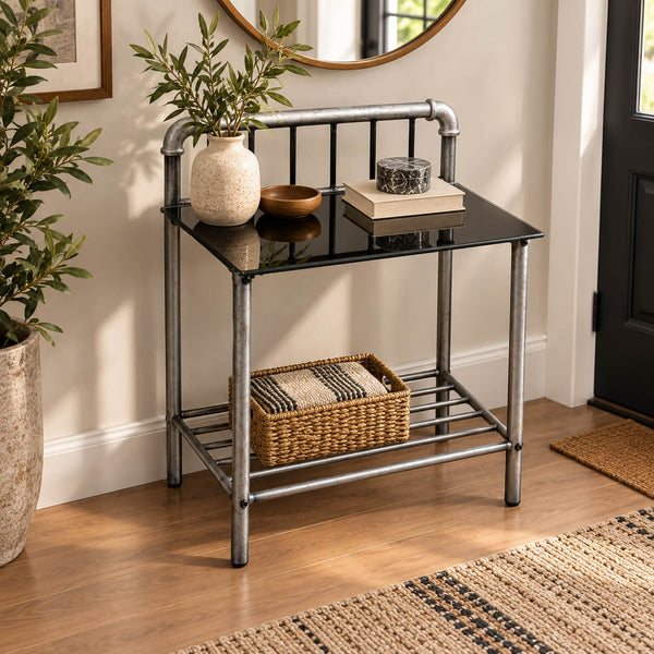 A small industrial-style entry table with a black glass top, silver metal frame, and an open lower shelf holding a woven basket. The table is styled with a vase of greenery, a bowl, and books in a bright room with wood flooring and a mirror.