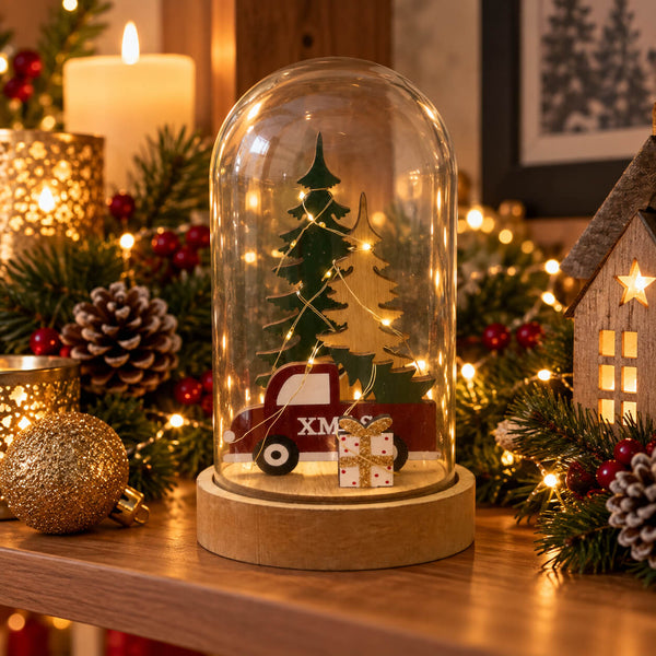 A Christmas-themed glass cloche dome on a round wooden base, decorated with miniature green and yellow Christmas trees, a red truck with 'XMAS' on the side, and a small wrapped gift. The cloche is displayed on a wooden shelf surrounded by holiday decorations including pine branches, pinecones, a lit candle, and warm string lights.