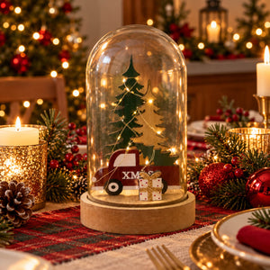 A Christmas-themed glass cloche centerpiece on a wooden base, featuring a miniature green Christmas tree, warm string lights, a red truck with 'XMAS' on the side, and a wrapped gift. The cloche sits on a decorated holiday table with candles, pine branches, and ornaments.
