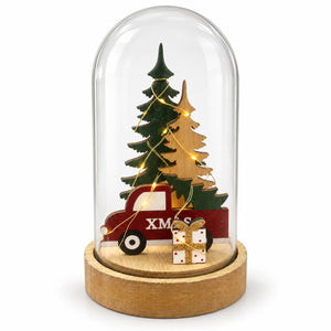 A decorative holiday dome display featuring a red pickup truck carrying two Christmas trees, wrapped in warm white fairy lights, with a small gift box in front, all enclosed in a clear glass dome on a round wooden base.