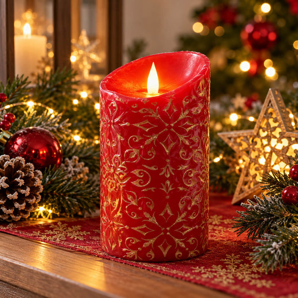 A red faux LED pillar candle with a gold snowflake and scroll pattern, lit with a warm flame, set on a wooden table surrounded by Christmas decorations including pine branches, ornaments, a pinecone, and a glowing star lantern.