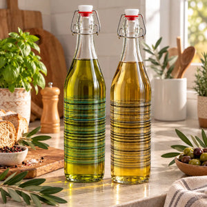Two tall clear glass swing-top bottles filled with olive oil, standing on a kitchen counter with bread, basil, olives, and kitchen items in the background.