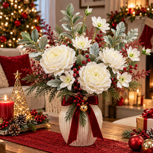 A festive Christmas floral arrangement in a ribbed white ceramic vase, featuring large white flowers, silver-green foliage, red berries, and a deep red ribbon bow with a small gold ornament, set on a decorated holiday table in a cozy living room.