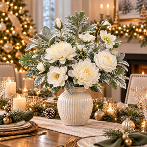 A festive holiday floral centerpiece in a ribbed cream vase, featuring white flowers and silver-dusted green foliage, displayed on a decorated Christmas dinner table.