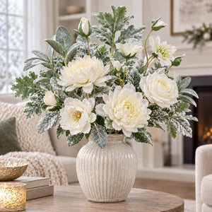 A faux floral arrangement of white flowers and green foliage with a textured cream vase, styled on a wooden table in a cozy living room setting.
