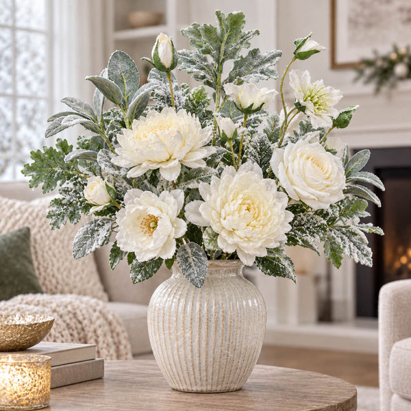 A faux floral arrangement of white flowers and green foliage with a textured cream vase, styled on a wooden table in a cozy living room setting.