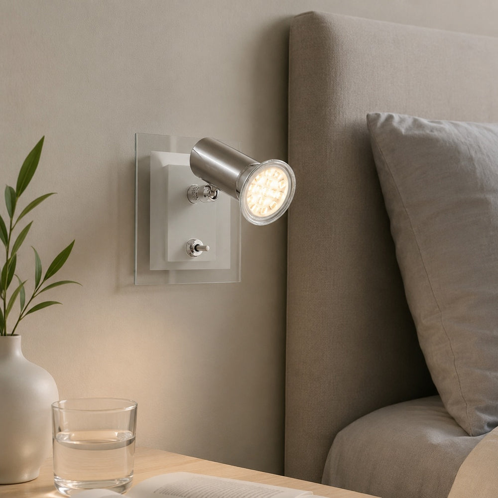 A modern wall-mounted spotlight lamp with a brushed silver metal head and clear acrylic backplate, shown lit in a bedroom setting next to a bed with a gray headboard.