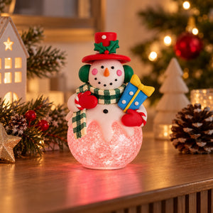 A festive snowman holiday light decoration sitting on a wooden table, surrounded by Christmas decorations like a lit house, pinecones, and a blurred Christmas tree in the background.