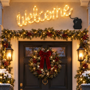 A festive holiday front door decorated with a glowing 'Welcome' sign above, a green garland with warm white lights and red berries, a wreath with a large red bow, and lanterns on both sides.