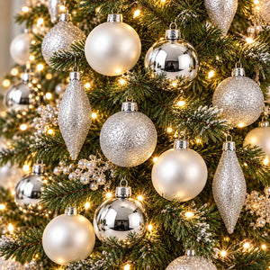 Silver and white Christmas tree ornaments hanging on a green pine tree with warm white string lights.