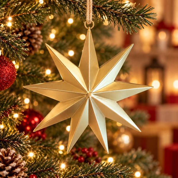 A metallic gold 12-point star Christmas ornament hanging from a tree branch with warm holiday lights and blurred ornaments in the background.