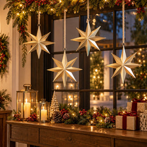 Four hanging metallic star ornaments with a warm white glow, displayed on a decorated Christmas windowsill with candles, pinecones, a small tree, and wrapped gifts.
