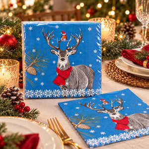 Blue holiday paper napkins featuring a gray reindeer with antlers, a red scarf, white snowflakes, pine branches, and a small Santa figure on the antlers, shown on a decorated dining table with candles, plates, and greenery.