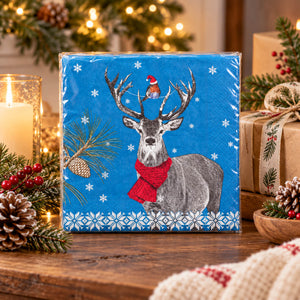 A square blue paper napkin with a festive Christmas design featuring a gray deer wearing a red scarf, a small red bird on the deer's antlers, white snowflakes, and pine branches, displayed on a wooden table with holiday decorations in the background.