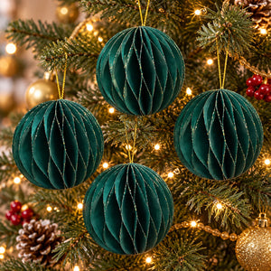 Four dark teal green honeycomb paper ball ornaments hanging on a decorated Christmas tree with warm string lights.