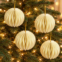 Four cream-colored honeycomb paper Christmas tree ornaments hanging on a green Christmas tree with warm string lights.