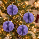 Four purple honeycomb paper Christmas tree ornaments hanging on a lit green Christmas tree with warm white lights and pinecones.