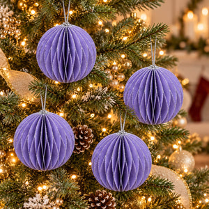 Four purple honeycomb paper Christmas tree ornaments hanging on a lit green Christmas tree with warm white lights and pinecones.