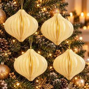 Four cream-colored, honeycomb-style paper Christmas ornaments hanging on a decorated Christmas tree with warm string lights, pinecones, and gold ornaments.