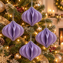 Four lavender purple honeycomb paper Christmas tree ornaments hanging on a decorated Christmas tree with warm lights and gold ornaments.