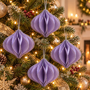 Four lavender purple honeycomb paper Christmas tree ornaments hanging on a decorated Christmas tree with warm lights and gold ornaments.